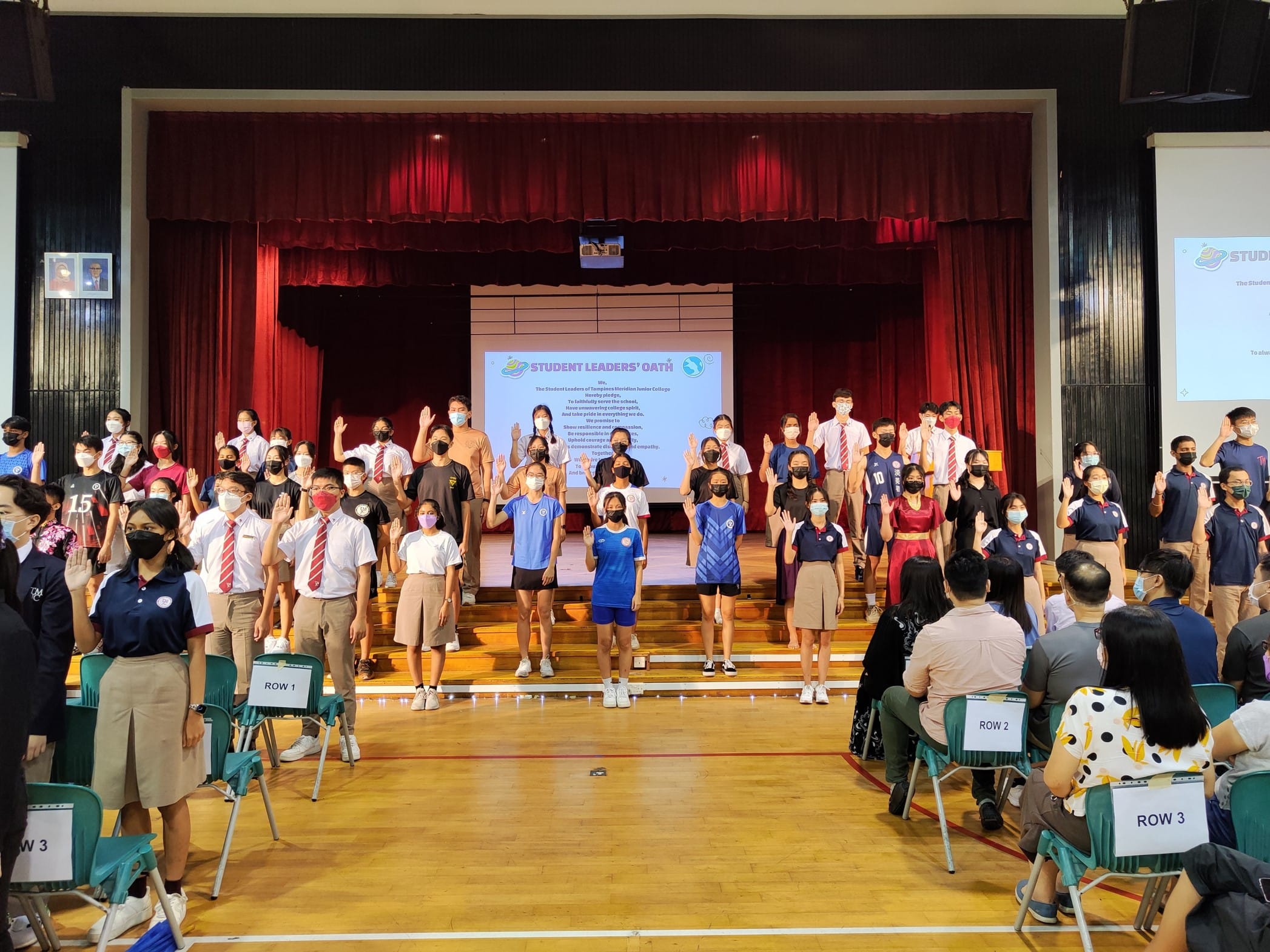 Students wearing masks raise their right hands during a Student Leaders' Oath ceremony in a school hall.